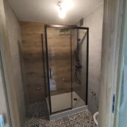 Nowoczesna kabina prysznicowa z czarnymi profilami, drewnopodobna ściana, wzorzysta podłoga, szare płytki na ścianach, widok zza framugi drzwi.