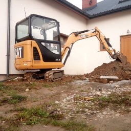 Mateusz Mostowik konserwacja i naprawa pojazdów samochodowych pit stop - Żółta mini koparka CAT 301.6 nabiera ziemię na tle białej ściany budynku z brązowymi drzwiami i oknem.