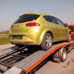 Mateusz Mostowik konserwacja i naprawa pojazdów samochodowych pit stop - Żółty samochód osobowy Seat Leon przewożony na pomarańczowej lawecie z metalową platformą, widok z tyłu na tle zielonego pola i błękitnego nieba.