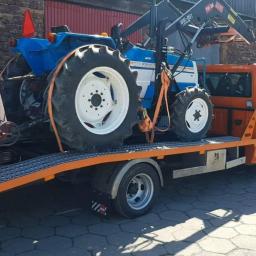 Mateusz Mostowik konserwacja i naprawa pojazdów samochodowych pit stop - Pomarańczowa laweta z niebieskim traktorem rolniczym marki Mitsubishi i podczepionymi bronami talerzowymi na platformie, zabezpieczonymi pomarańczowymi pasami transportowymi na tle ceglanego...