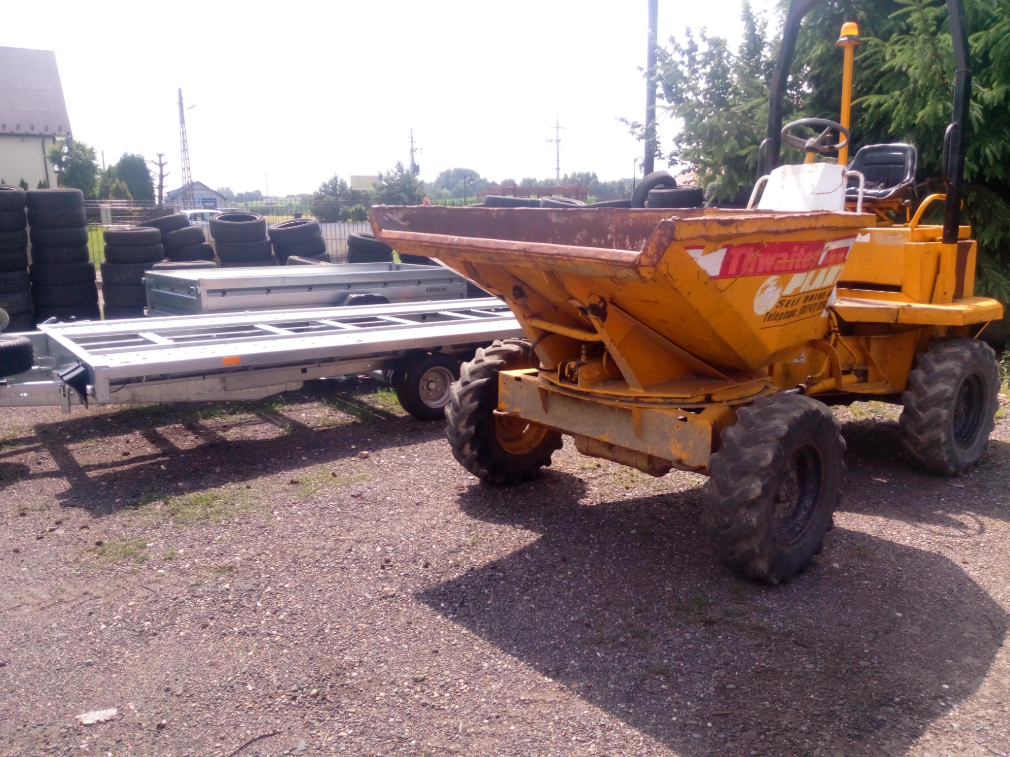 Żółta wywrotka budowlana marki Thwaites na żwirowym podłożu, z przyczepą i stosem opon w tle.