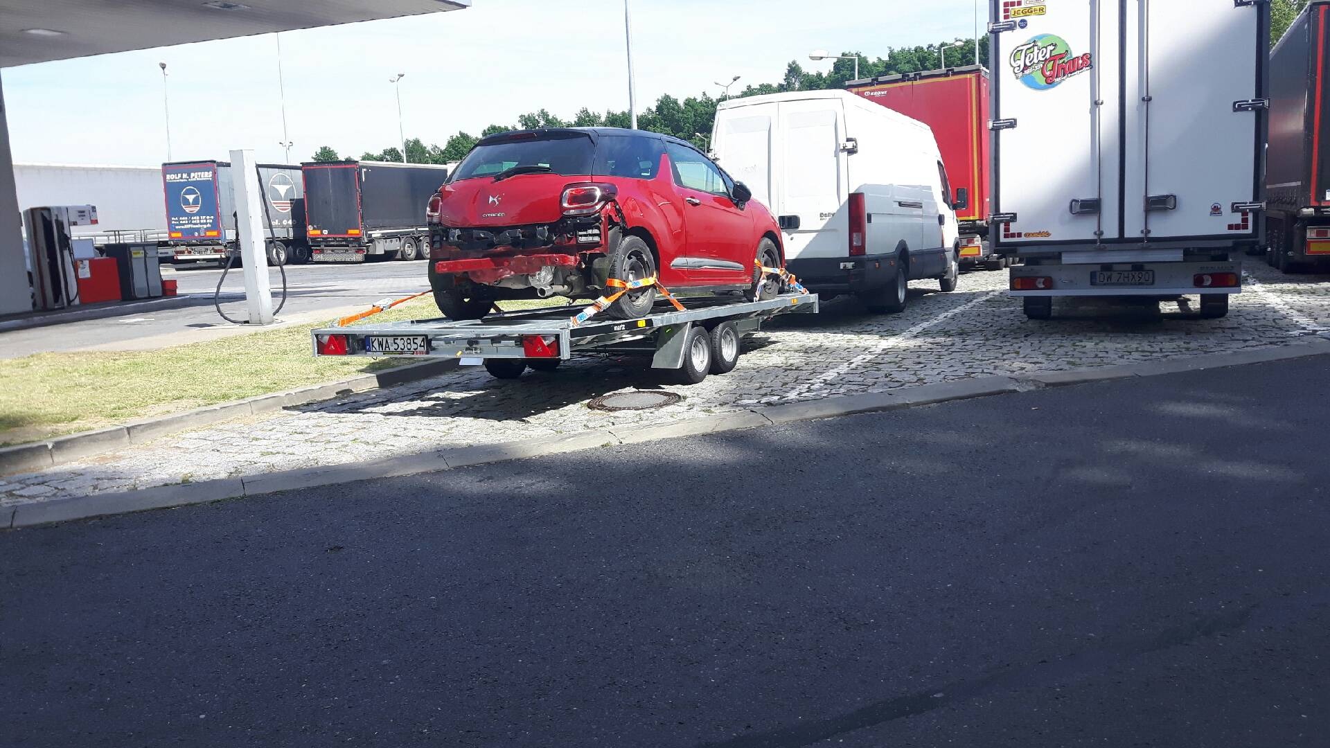 Uszkodzony czerwony samochód Citroen DS3, zabezpieczony pomarańczowymi pasami, transportowany na dwuosiowej przyczepie lawetą na parkingu stacji benzynowej, z widocznymi ciężarówkami w tle.