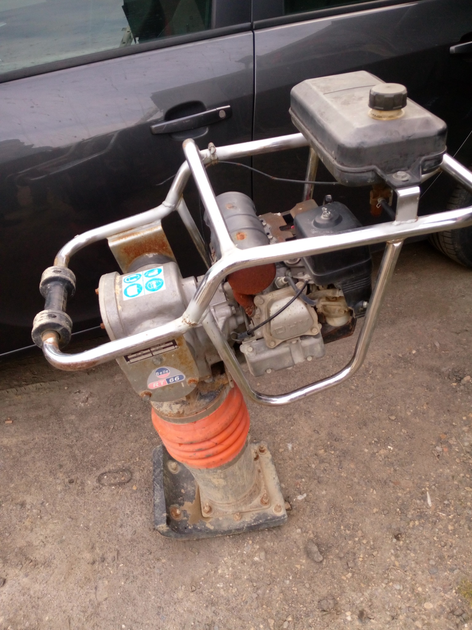 Używana ubijarka wibracyjna Belle RT66, z pomarańczowym mieszkiem i srebrną ramą, stoi na ziemi obok ciemnego samochodu.