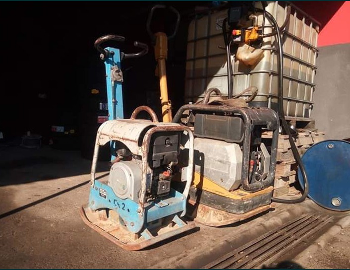 Dwie ubijarki wibracyjne (zagęszczarki) z silnikami spalinowymi, jedna niebieska, druga czarna, stoją na brudnej betonowej posadzce w warsztacie, obok zbiornik IBC na palecie i niebieska beczka.