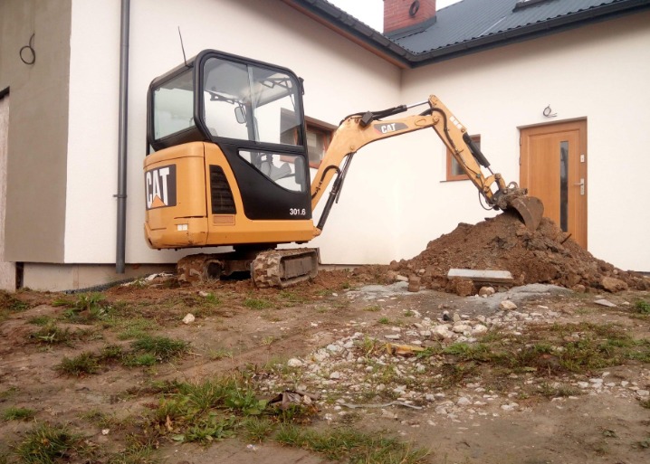 Żółta mini koparka CAT 301.6 nabiera ziemię na tle białej ściany budynku z brązowymi drzwiami i oknem.