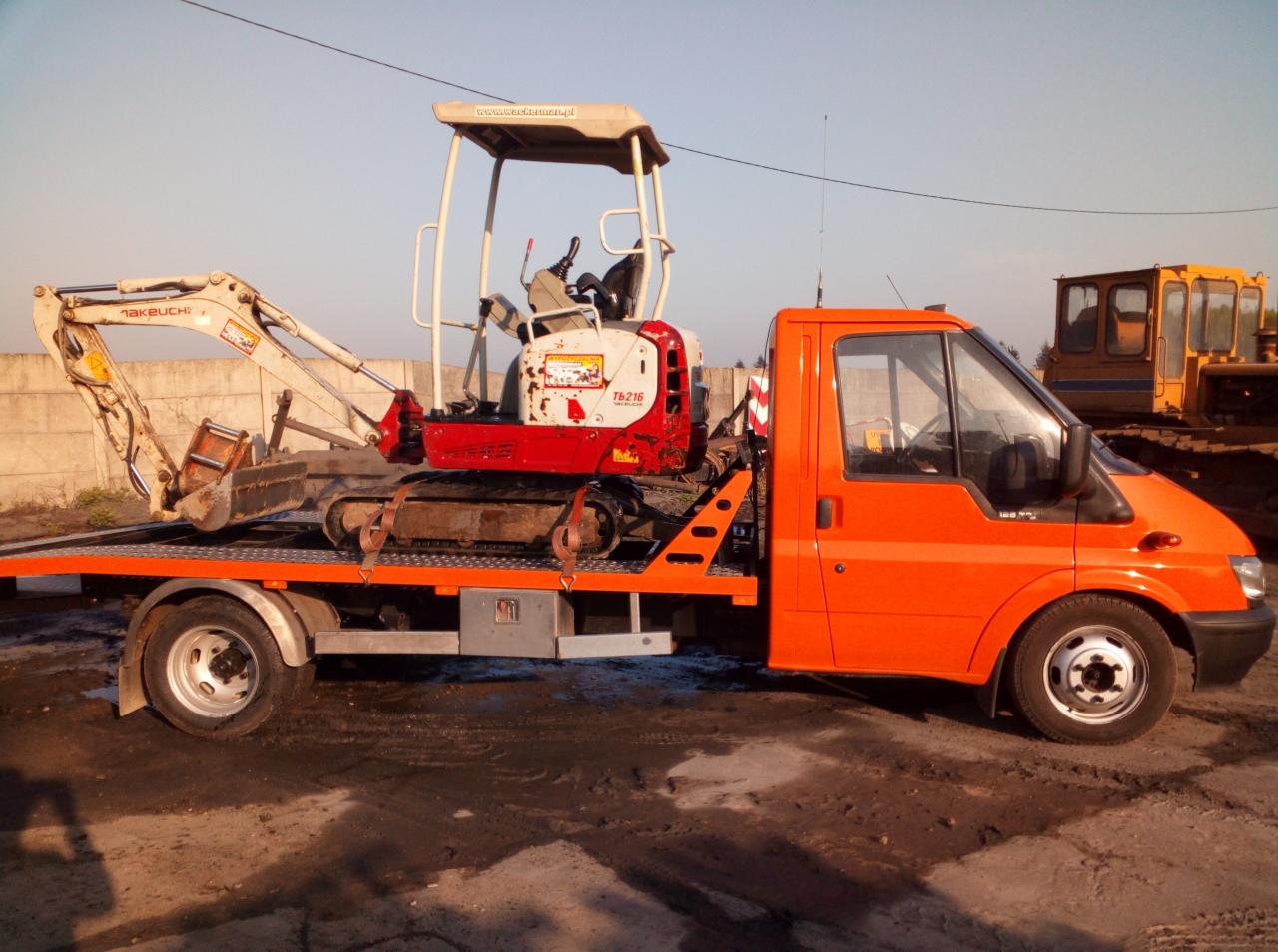 Pomarańczowa laweta z mini koparką Takeuchi TB216 na platformie, w tle żółta koparka gąsienicowa i betonowy płot.