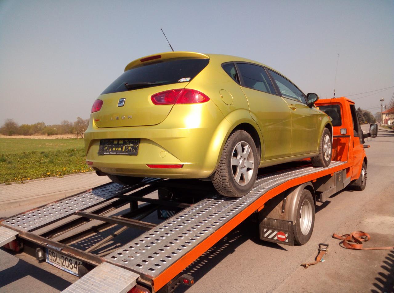 Żółty samochód osobowy Seat Leon przewożony na pomarańczowej lawecie z metalową platformą, widok z tyłu na tle zielonego pola i błękitnego nieba.