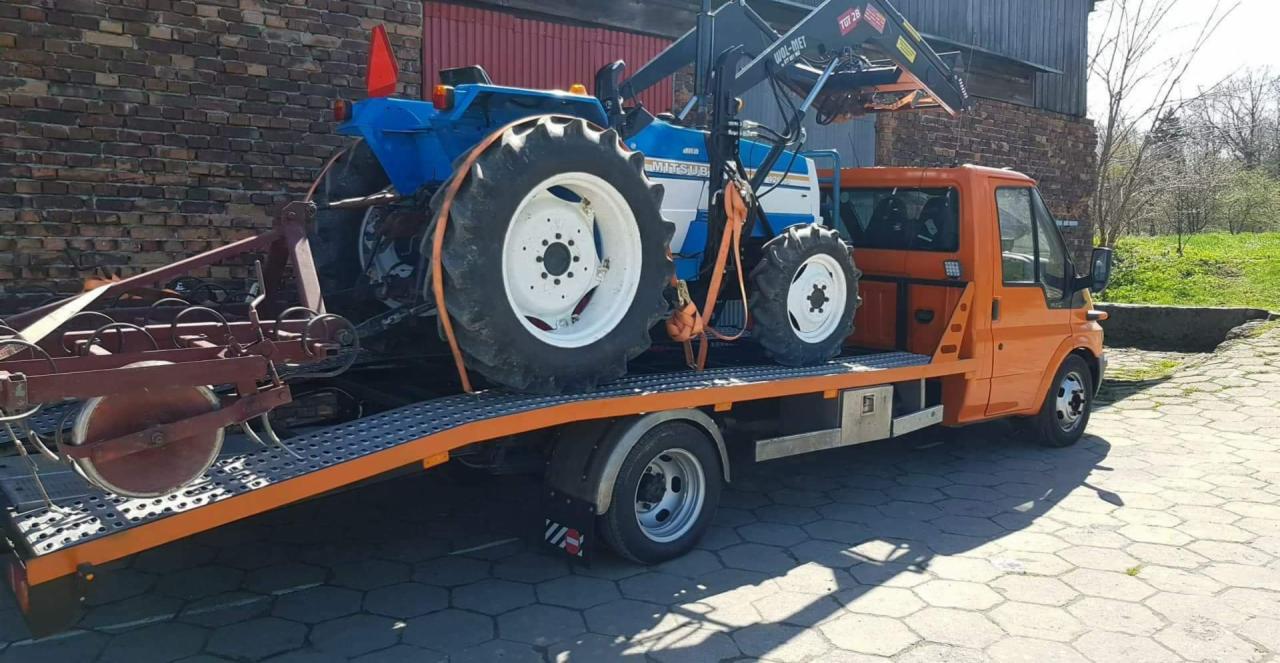 Pomarańczowa laweta z niebieskim traktorem rolniczym marki Mitsubishi i podczepionymi bronami talerzowymi na platformie, zabezpieczonymi pomarańczowymi pasami transportowymi na tle ceglanego...