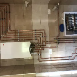 Widok z góry na kotłownię z piecem, rozdzielaczem i miedzianymi rurami instalacji grzewczej zamocowanymi do kafelkowanej ściany.