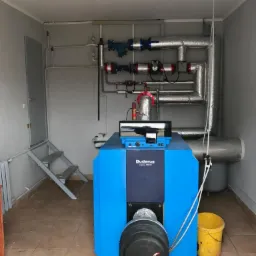 Niebieski piec grzewczy Buderus Logano G315 w kotłowni z widocznymi rurami instalacji, schodkami i żółtym wiadrem.