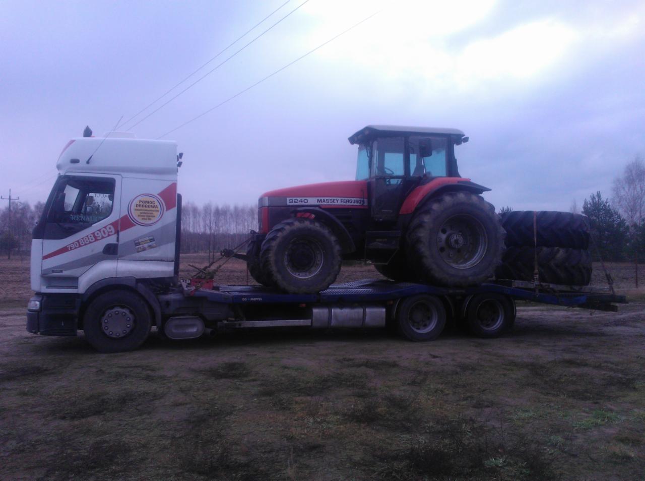 Ciężarówka Renault z platformą przewozi traktor Massey Ferguson 9240 i opony na tle pochmurnego nieba i drzew. Widoczny numer telefonu na kabinie.