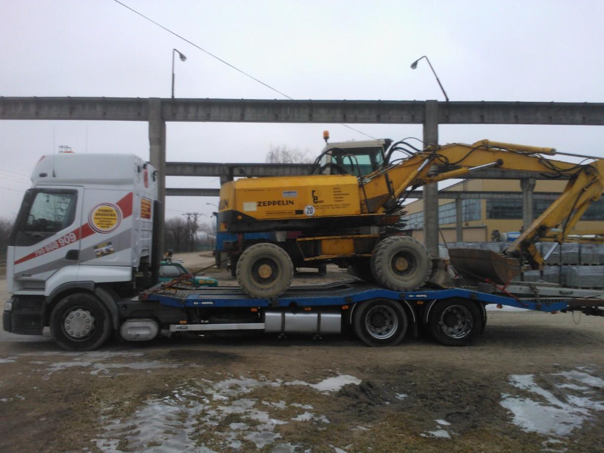 Laweta z żółtą koparką Zeppelin na niebieskiej platformie, transportowana przez białą ciężarówkę z numerem telefonu na tle szarego nieba.