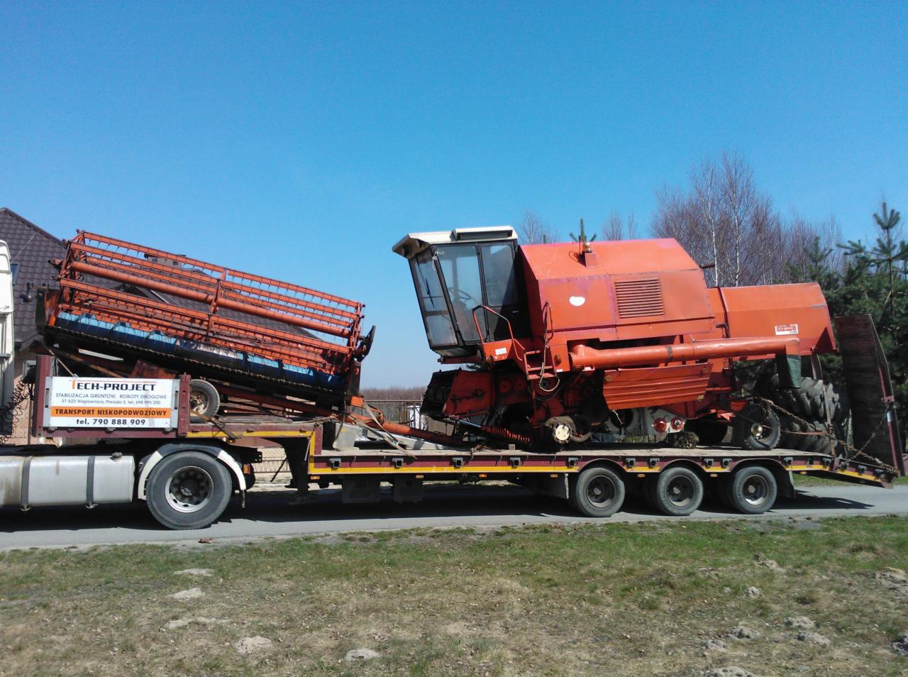 Ciężarówka z lawetą transportuje kombajn rolniczy. Kombajn koloru czerwonego. Na lawecie widoczny również element tnący. Błękitne niebo w tle.