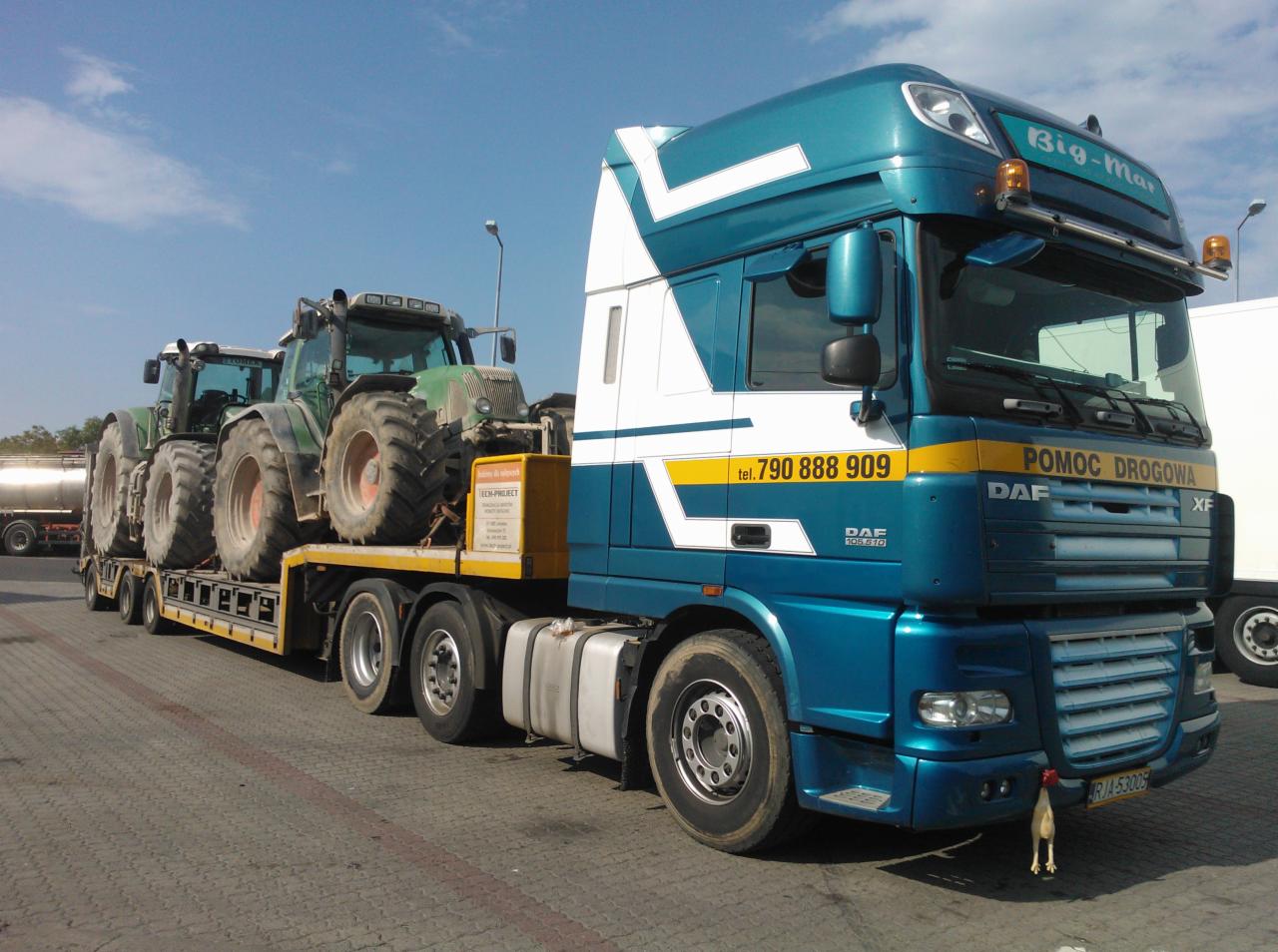 Laweta DAF XF przewozi dwa zielone traktory Fendt. Widoczny numer telefonu i napis POMOC DROGOWA. Nietypowa ozdoba na zderzaku.