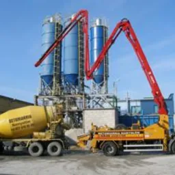 Żółta betonomieszarka podłączona do pompy do betonu na tle silosów i błękitnego nieba. Widoczny fragment budynku z lewej strony.