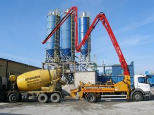 Żółta betonomieszarka podłączona do pompy do betonu na tle silosów i błękitnego nieba. Widoczny fragment budynku z lewej strony.