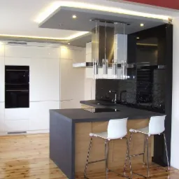 Nowoczesna kuchnia z wyspą i drewnianym blatem, oświetlenie LED podsufitowe, czarne szkło na ścianie, drewniana podłoga i dwa białe hokery.