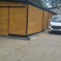 FAR-BUD Tomasz Cieślar - Nowoczesna, drewniana konstrukcja z przesuwnymi drzwiami, częściowo zasłonięta przez pomarańczową ciężarówkę z napisem 'Kamień Dekoracyjny', biały van z otwartymi drzwiami na kostce brukowej.