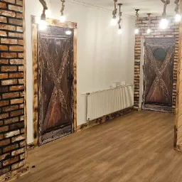 Wnętrze pokoju z drewnianą podłogą, ceglaną ścianą, rustykalnymi drzwiami z motywem stodoły i oświetleniem z żarówek wiszących na sznurach