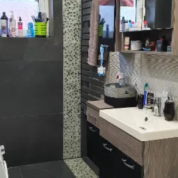 Nowoczesna łazienka z ciemnymi płytkami, mozaiką akcentującą i drewnopodobną zabudową umywalki z czarnymi frontami, widok na okno z roletą i dekoracyjnymi przedmiotami na parapecie.