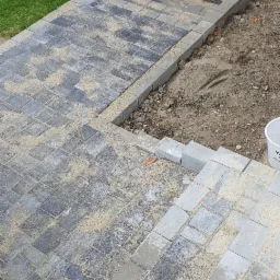 Ułożona kostka brukowa w odcieniach szarości prowadząca do drzwi wejściowych, posypana piaskiem kwarcowym, widoczny fragment trawnika i ściana budynku.