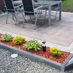 Szary stół ogrodowy z czterema krzesłami na tarasie z płytek imitujących drewno, obok rabata z korą i niskimi roślinami ozdobnymi, otoczenie wysypane drobnym kamieniem.