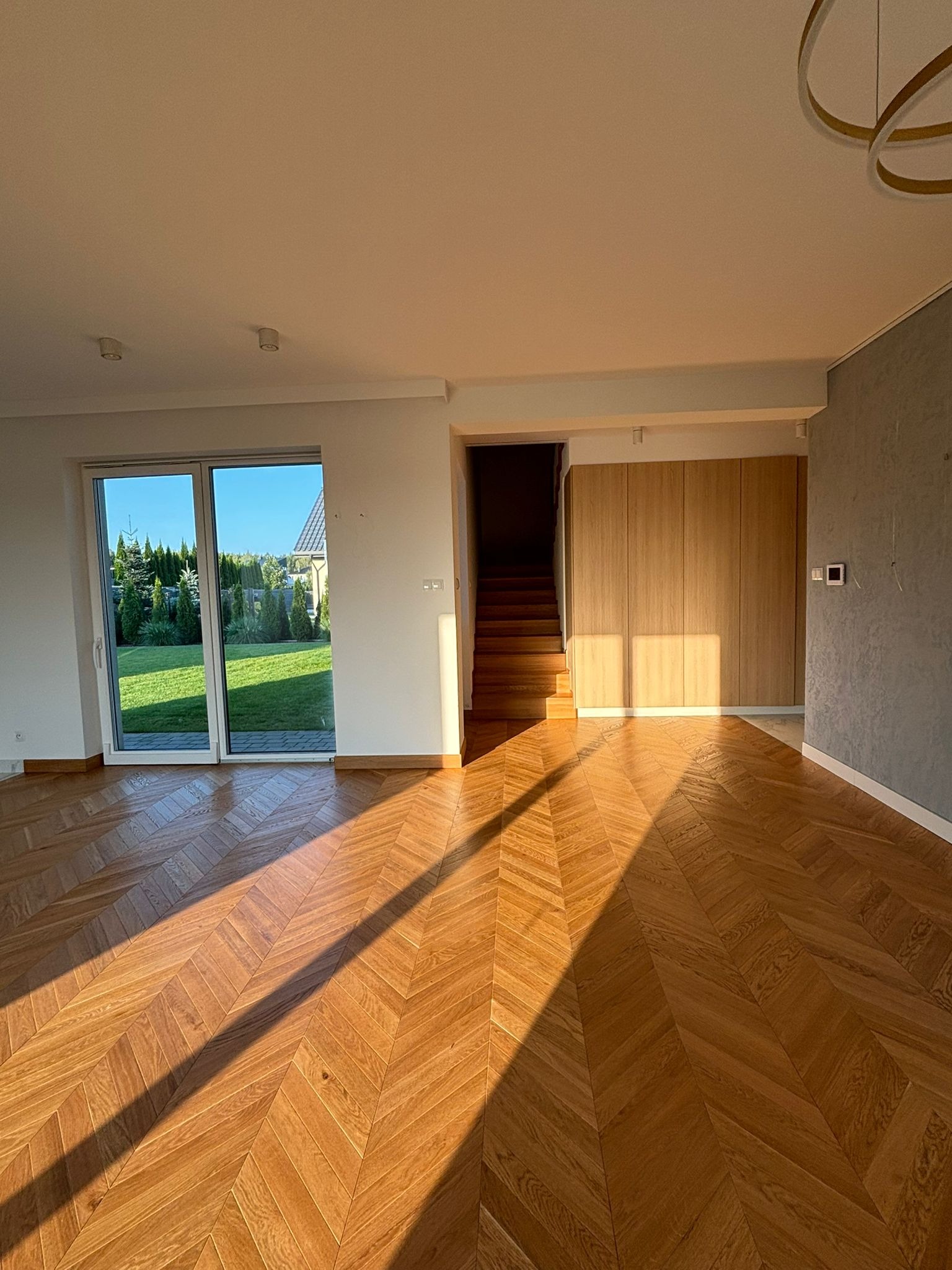 Jasne, przestronne wnętrze domu z parkietem ułożonym we wzór jodełki, duże okno z widokiem na ogród, schody i zabudowa meblowa z jasnego drewna, ściana wykończona w szarym tynku dekoracyjnym...