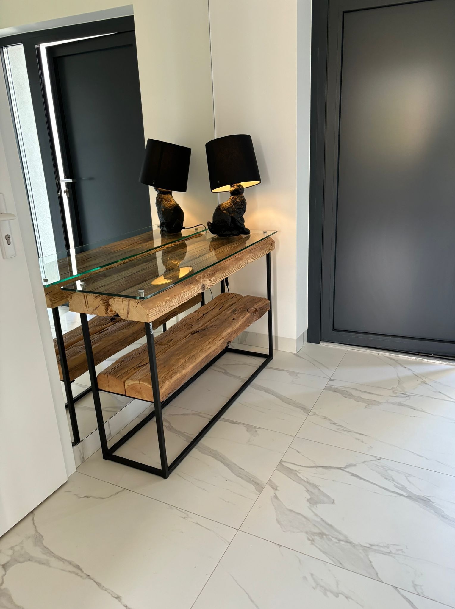 Nowoczesna konsola z blatem ze szkła i podstawą z surowego drewna, oświetlona dwiema lampkami z czarnymi abażurami w kształcie kotów, na tle białej ściany i grafitowych drzwi, na podłodze z białego...