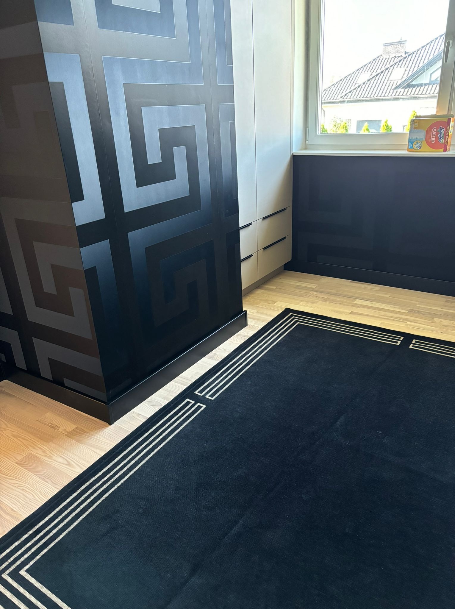 Pokój po odświeżeniu: czarny wzorzysty panel ścienny, beżowa szafa wnękowa, ciemny dywan z jasnymi paskami na parkiecie, okno z widokiem na dom.