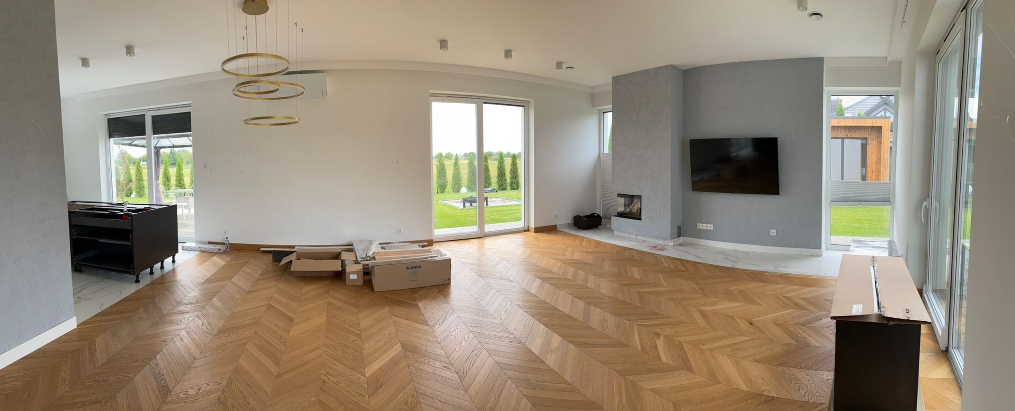 Salon w trakcie wykańczania: parkiet w jodełkę, szary kominek z telewizorem, złote lampy pierścieniowe, częściowo rozpakowane meble kuchenne, widok na ogród przez duże okna.