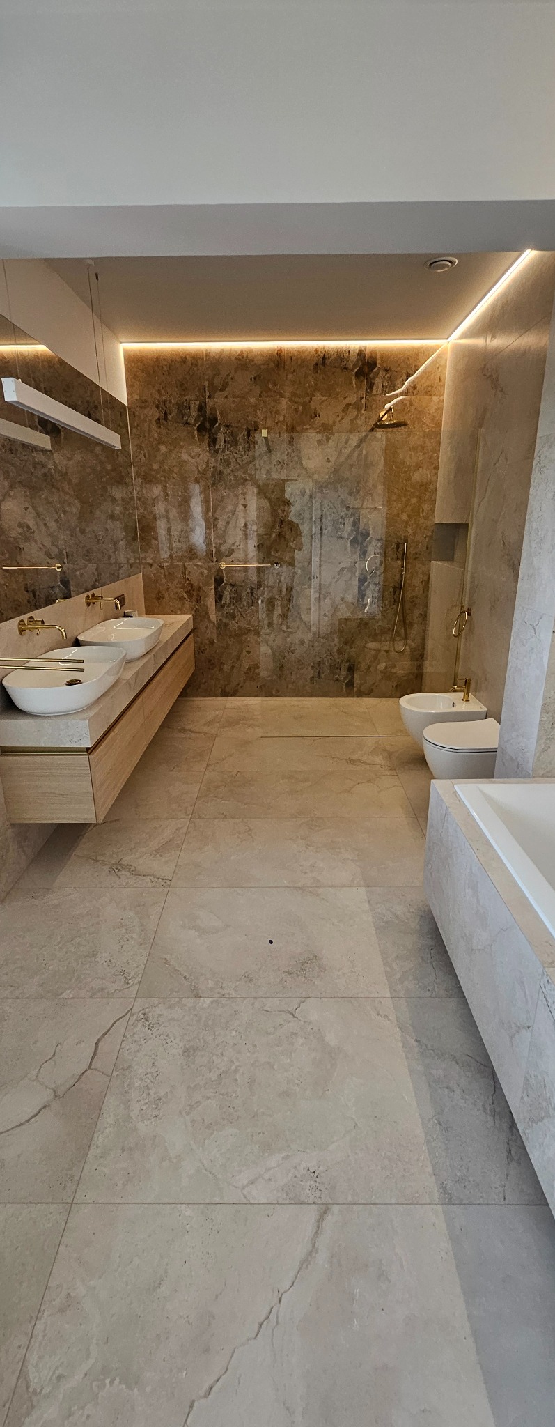 Elegancka, wykończona łazienka w Wiśle z marmurowymi płytkami, oświetleniem LED i nowoczesną armaturą w kolorze złota, obejmująca umywalki, bidet, toaletę i wannę.