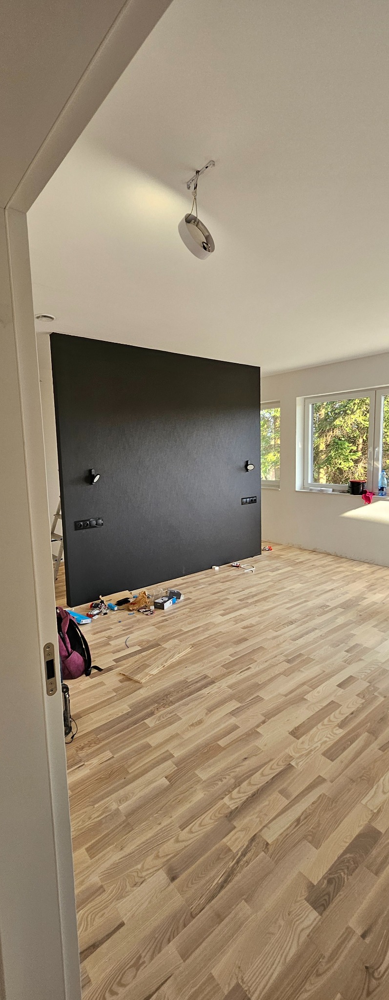 Wykończony pokój z parkietem ułożonym w jodełkę, czarna ściana akcentująca z zamontowanymi kinkietami i gniazdkami, widoczne narzędzia i akcesoria po remoncie, okno z widokiem na zieleń.