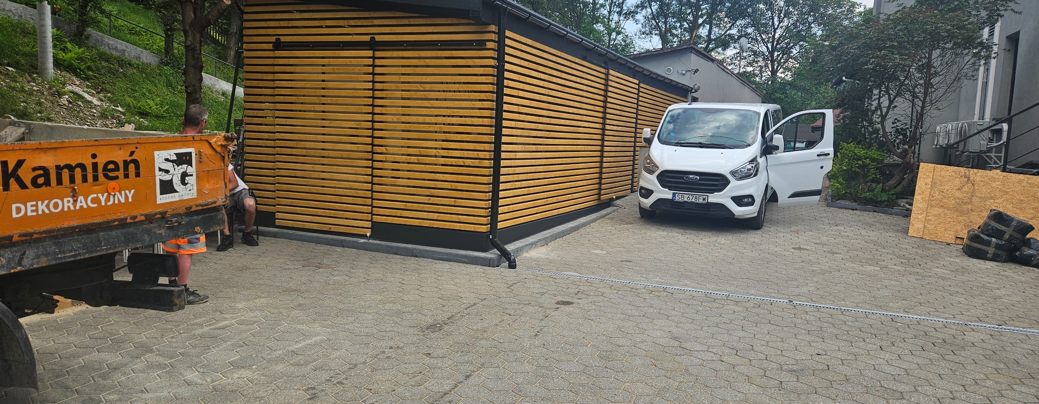 Nowoczesna, drewniana konstrukcja z przesuwnymi drzwiami, częściowo zasłonięta przez pomarańczową ciężarówkę z napisem 'Kamień Dekoracyjny', biały van z otwartymi drzwiami na kostce brukowej.