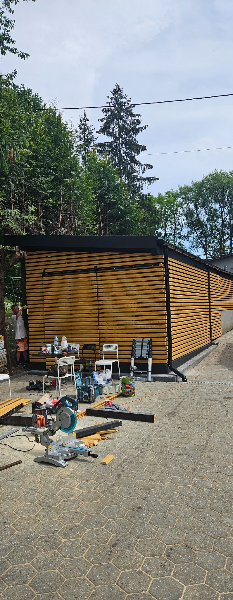 Nowoczesna, drewniana zabudowa tarasu z poziomymi lamelami w kolorze jasnego drewna, czarnym dachem i obramowaniem, w trakcie montażu, z widocznymi narzędziami i materiałami budowlanymi...