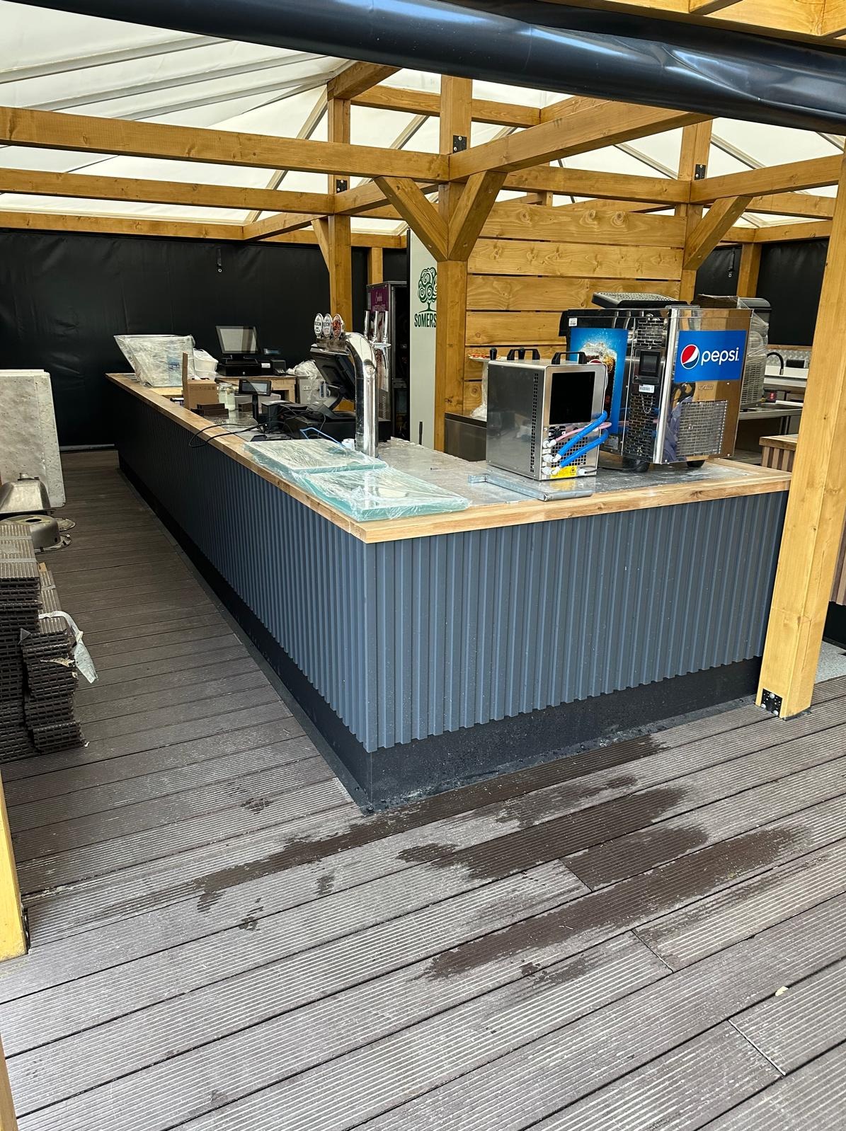 Drewniana konstrukcja z barem z szarymi panelami pionowymi, drewnianym blatem i sprzętem barowym, częściowo przykrytym folią ochronną, na zewnątrz na drewnianym tarasie z widocznymi plamami wody.