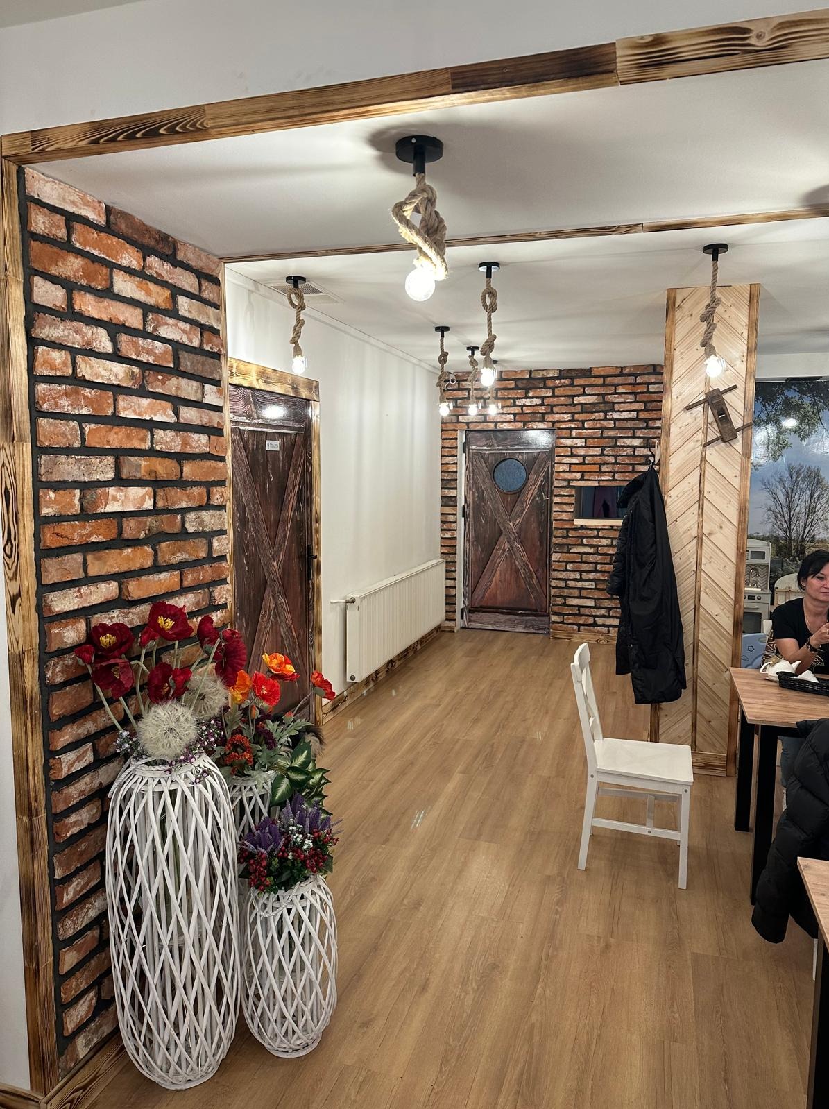 Wnętrze lokalu z dekoracyjną ścianą z cegły, drewnianymi detalami i lampami ze sznura, podłoga wyłożona panelami imitującymi drewno, w tle widoczni klienci przy stolikach.