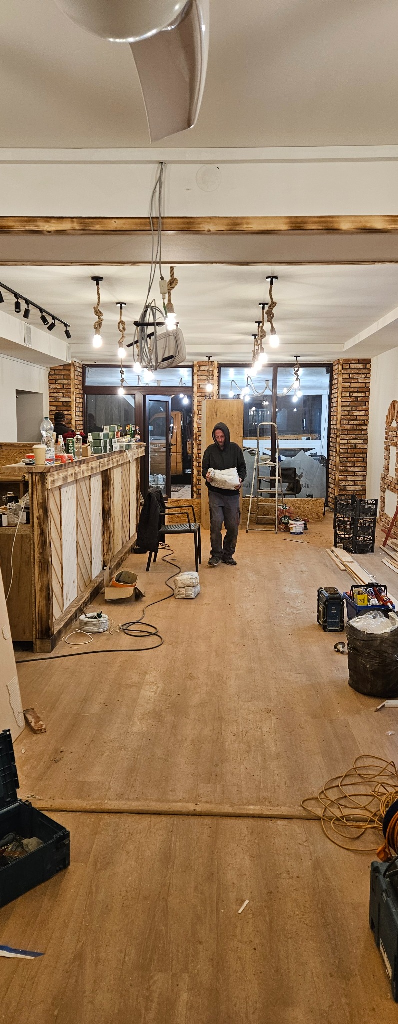 Wnętrze lokalu w trakcie prac wykończeniowych: drewniany bar z oświetleniem, widoczne narzędzia, mężczyzna w bluzie z kapturem przenoszący materiały, surowe ściany z elementami cegły, podłoga...
