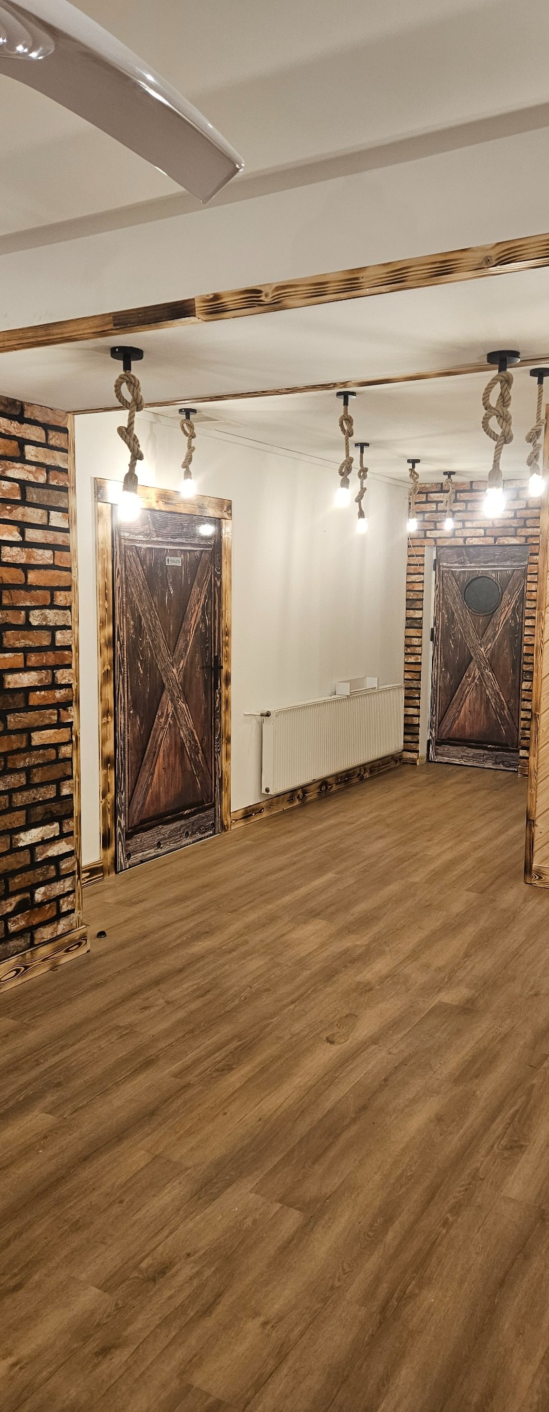 Wnętrze pokoju z drewnianą podłogą, ceglaną ścianą, rustykalnymi drzwiami z motywem stodoły i oświetleniem z żarówek wiszących na sznurach