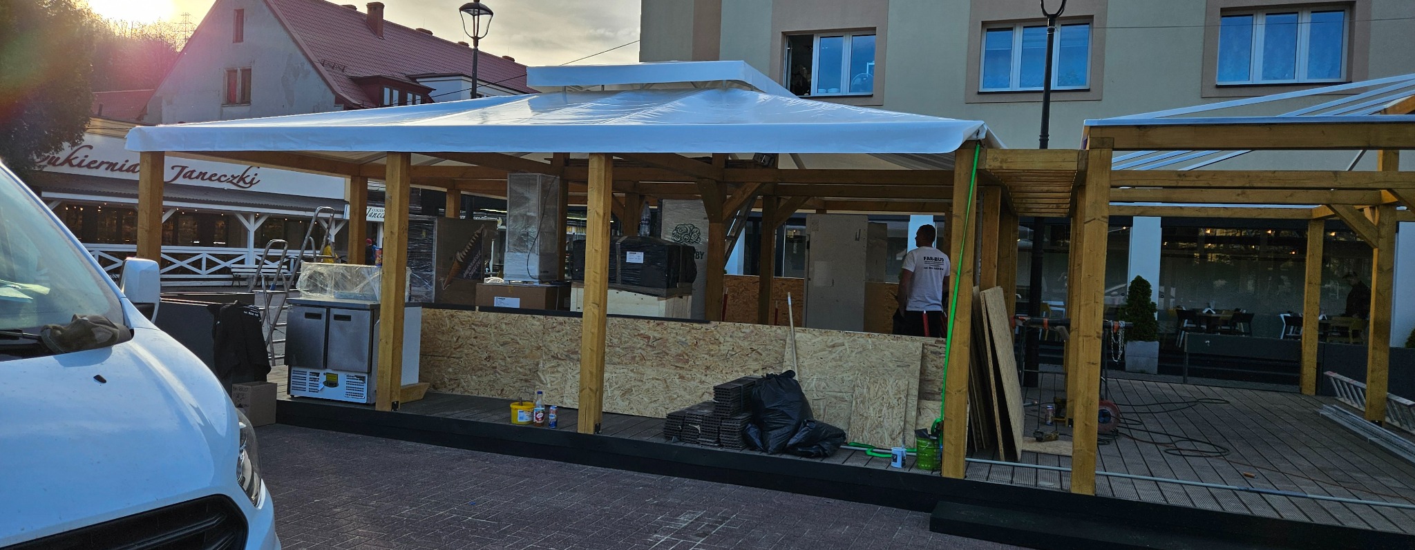 Konstrukcja drewniana z zadaszeniem z tworzywa sztucznego przed budynkiem, prawdopodobnie w trakcie montażu, widoczny pracownik w koszulce firmowej, elementy wyposażenia zapakowane w folie, płyty...