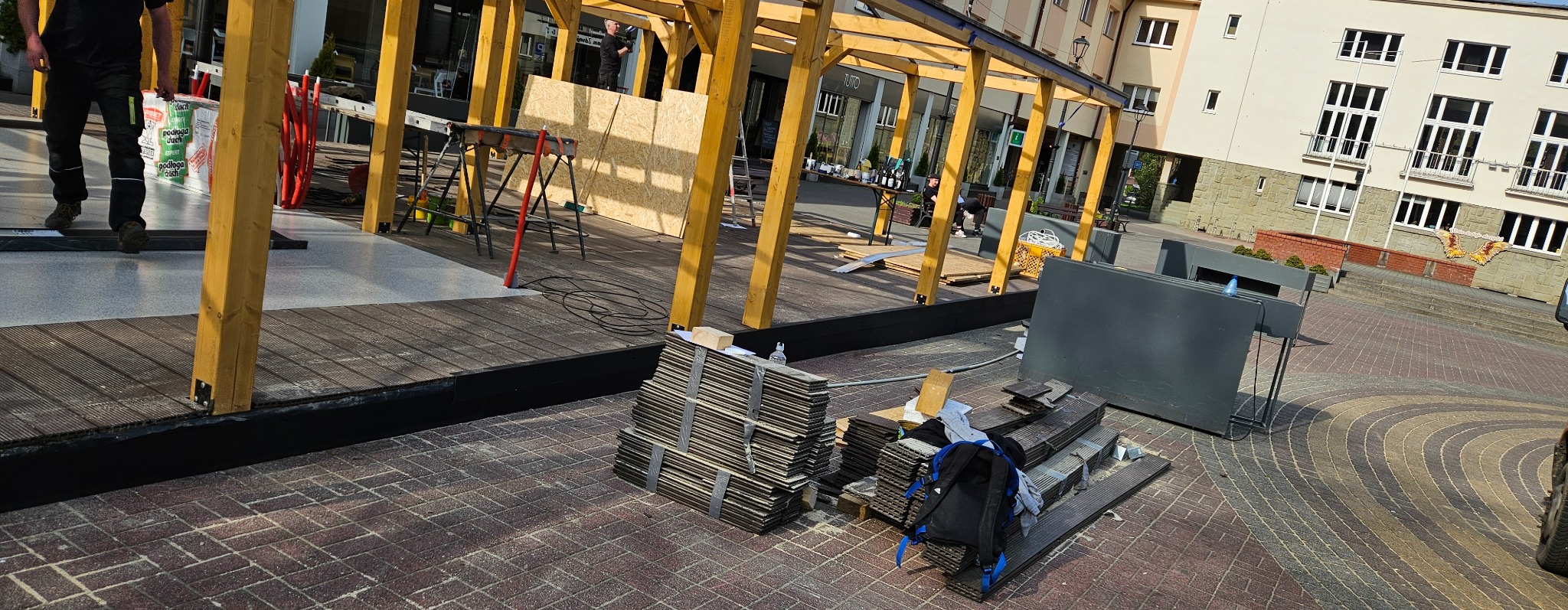 Konstrukcja drewniana z elementami płyty OSB, prawdopodobnie zadaszenie, w trakcie budowy na zewnątrz, widoczne narzędzia, materiały budowlane i pracownicy w tle.