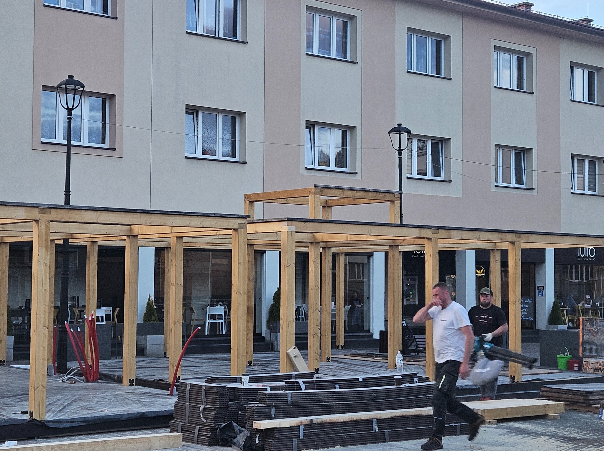 Konstrukcja drewniana zadaszenia w trakcie montażu na placu miejskim, widoczne elementy konstrukcyjne, narzędzia i pracownicy w tle budynku z oknami.