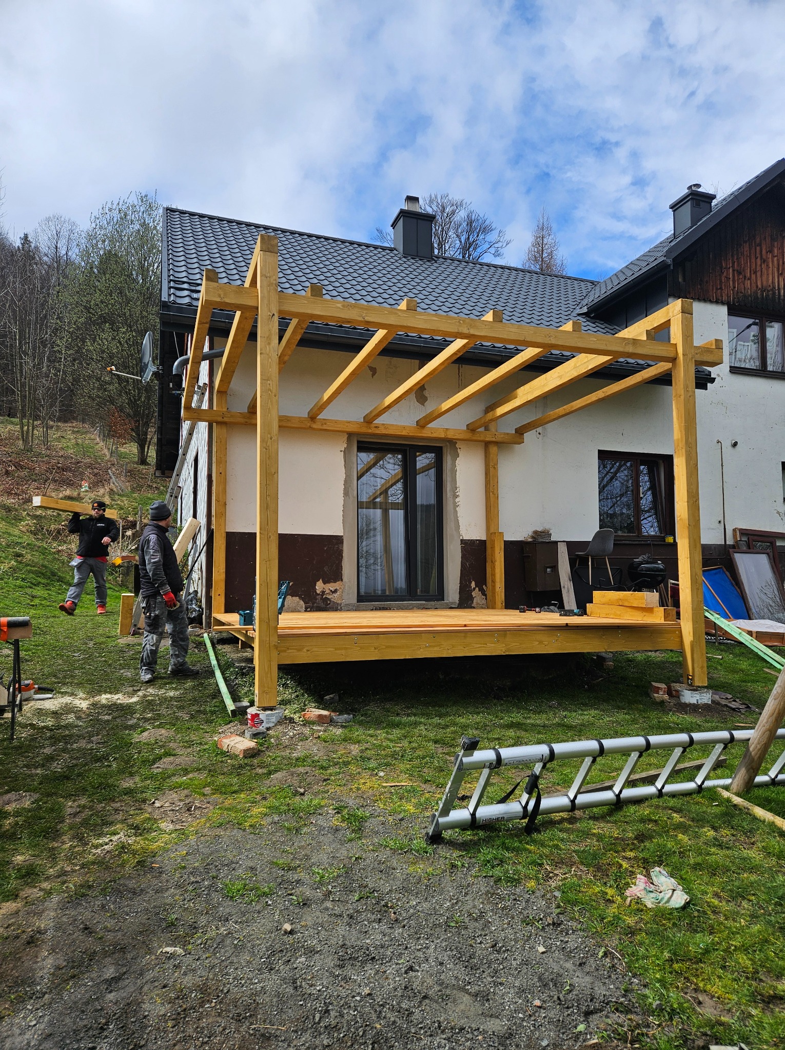 Budowa drewnianej konstrukcji zadaszenia tarasu przy domu jednorodzinnym, widoczni dwaj pracownicy w trakcie montażu belek, drabina oparta o konstrukcję, narzędzia na podłodze tarasu.