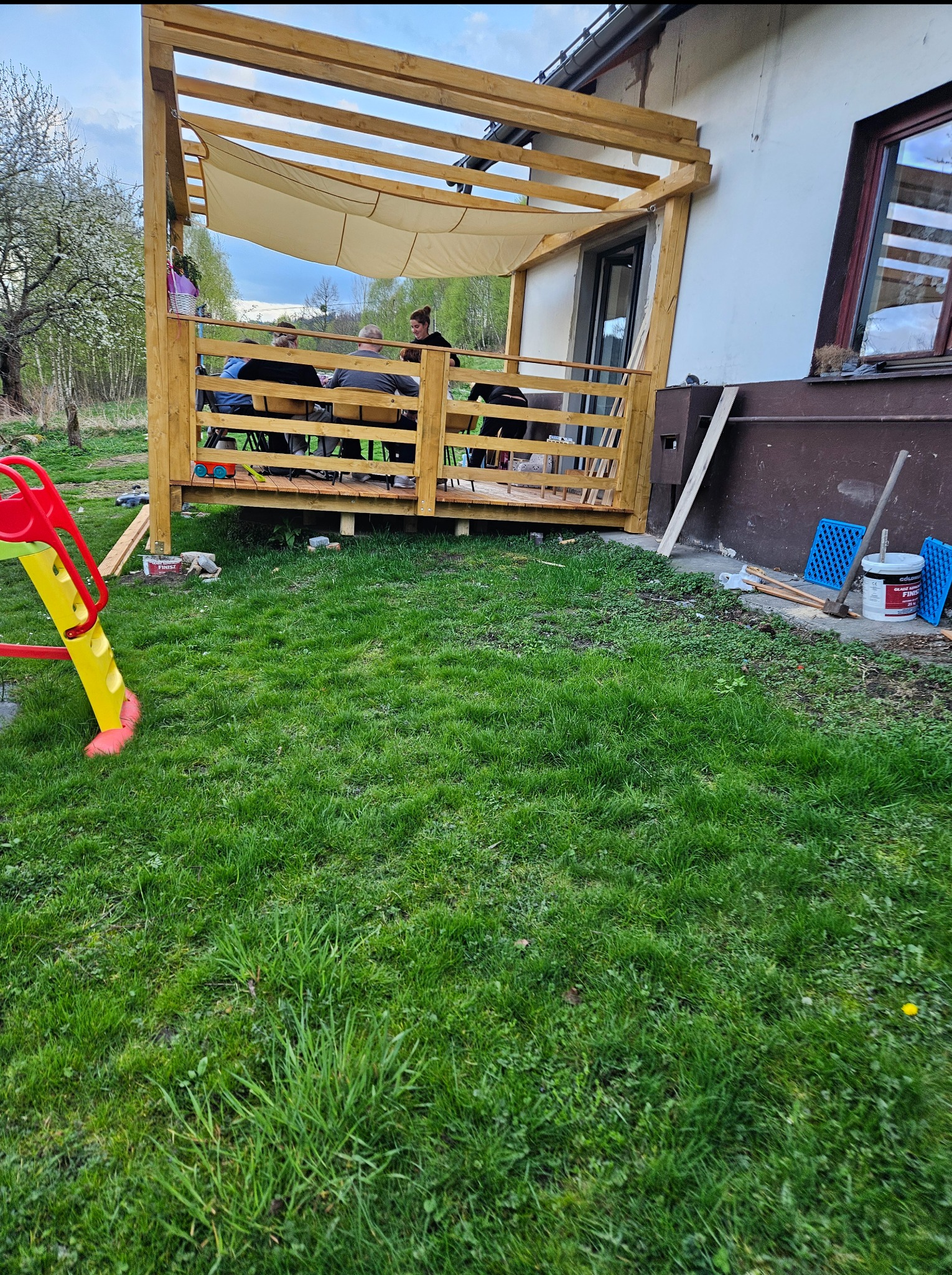Drewniana pergola przylegająca do budynku, z rozciągniętym materiałem jako zadaszenie, użytkowana przez osoby siedzące przy stole, widok z zewnątrz na trawnik i elementy placu zabaw.