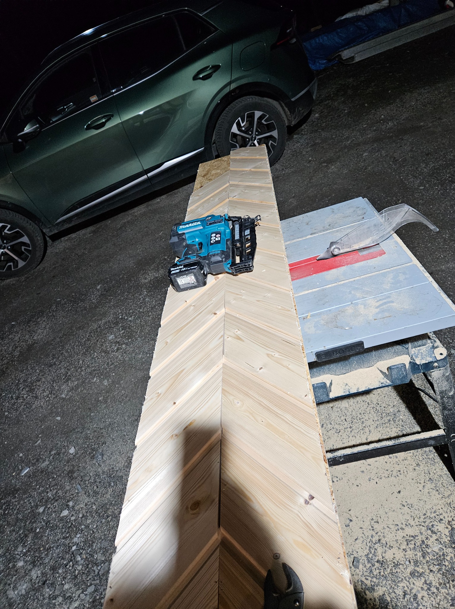 Deska z jasnego drewna ułożona w jodełkę na stole z piłą tarczową, obok leży akumulatorowa gwoździarka Makita; w tle widać zaparkowany samochód.
