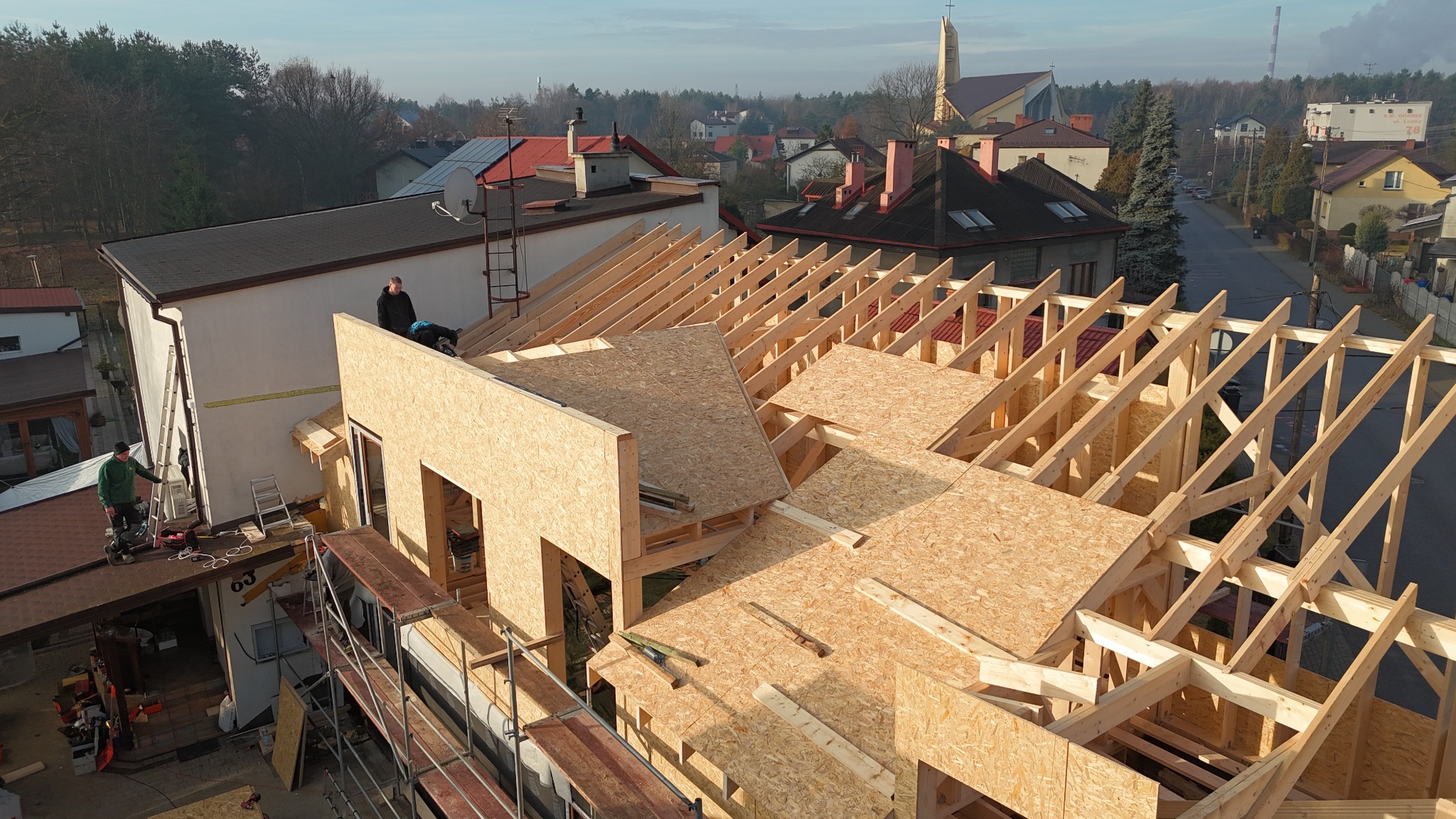 Konstrukcja dachu z drewnianych belek i płyt OSB w trakcie budowy domu jednorodzinnego, widok z góry na tle zabudowy mieszkalnej i kościoła.
