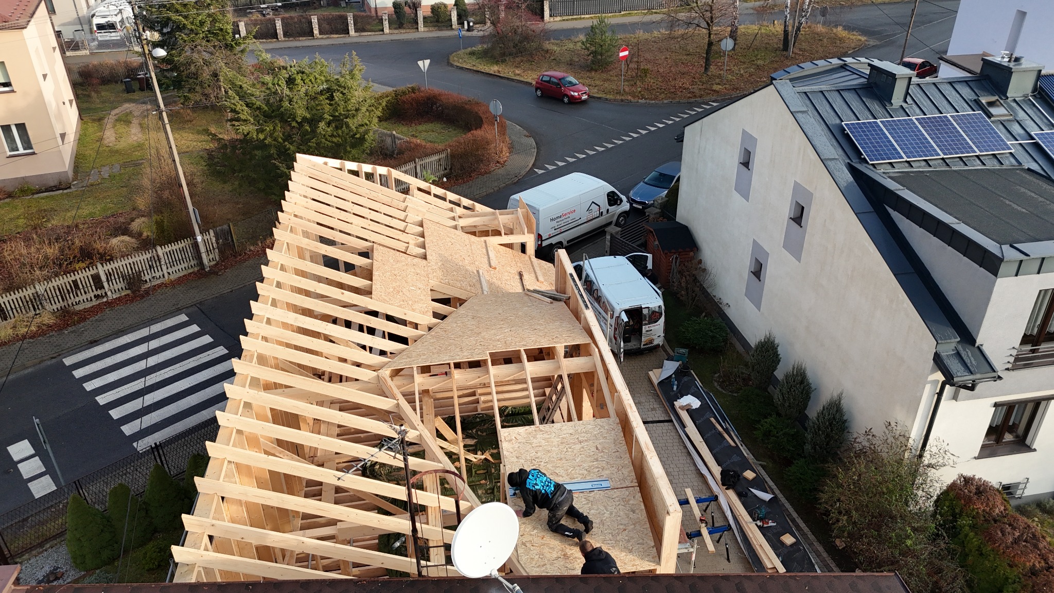 Konstrukcja drewniana dachu w trakcie budowy, widok z góry. Pracownicy montują elementy, w tle zabudowa miejska i zaparkowane busy.