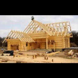 Surowy drewniany dom w trakcie budowy, widoczne elementy konstrukcyjne dachu i ścian, otoczenie placu budowy z porozrzucanymi deskami.