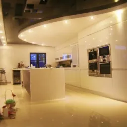 Nowoczesna, minimalistyczna kuchnia z białymi, lakierowanymi frontami, wyspą kuchenną i błyszczącą, odbijającą światło podłogą. Sufit podwieszany z oświetleniem punktowym.