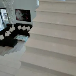 Nowoczesne, jasne schody prowadzące do salonu z ciemną sofą i kryształowym żyrandolem.