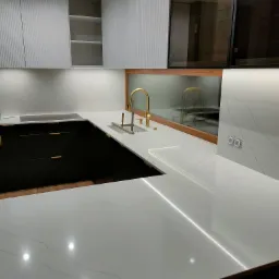 Biały blat kamienny z szarymi żyłkami w nowoczesnej kuchni z czarnymi szafkami i złotą armaturą, podświetlony listwą LED, widok na gładkie, białe fronty szafek górnych.